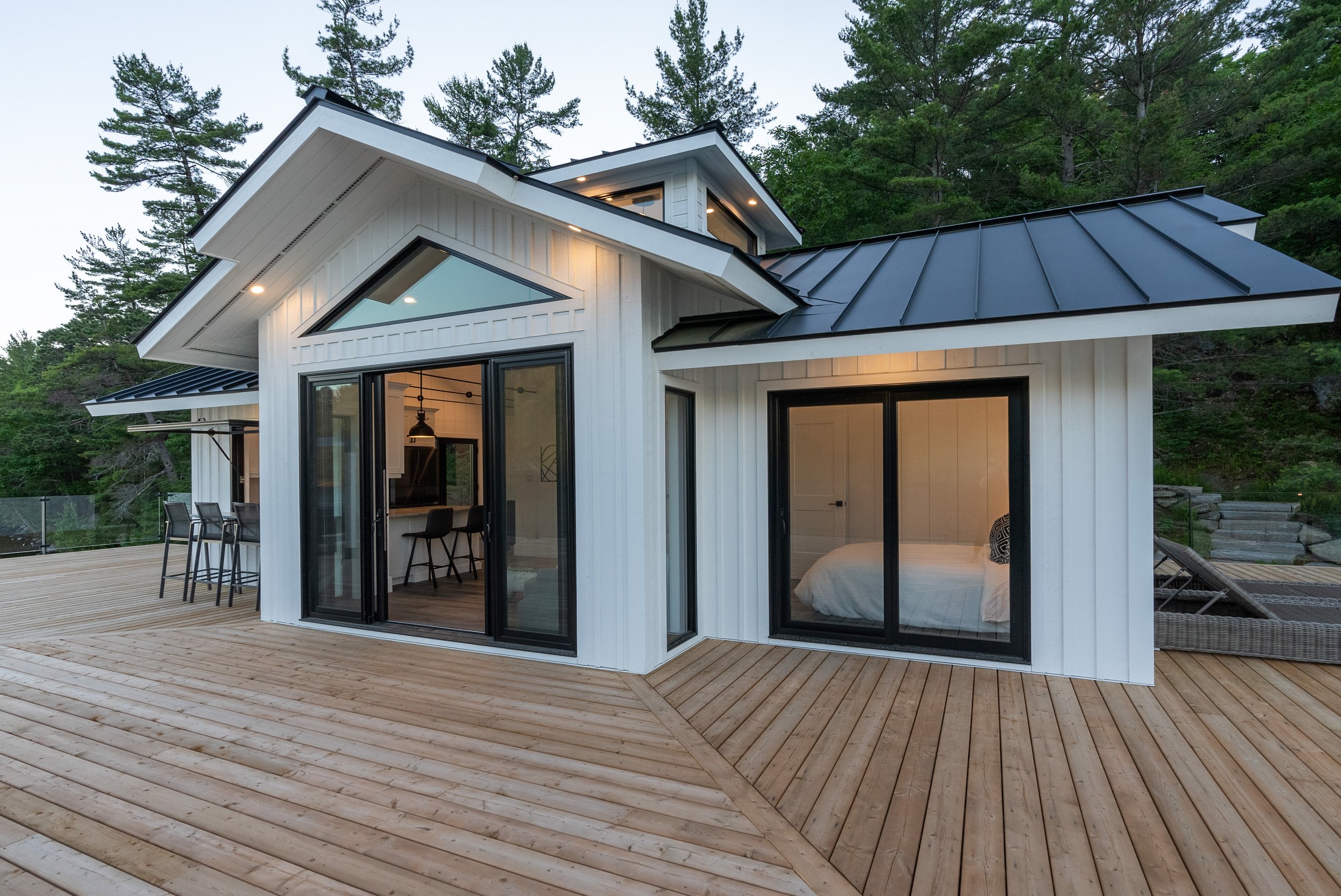 Modern house with white exterior and black roof, large glass windows, situated on a wooden deck with outdoor seating, surrounded by green trees.