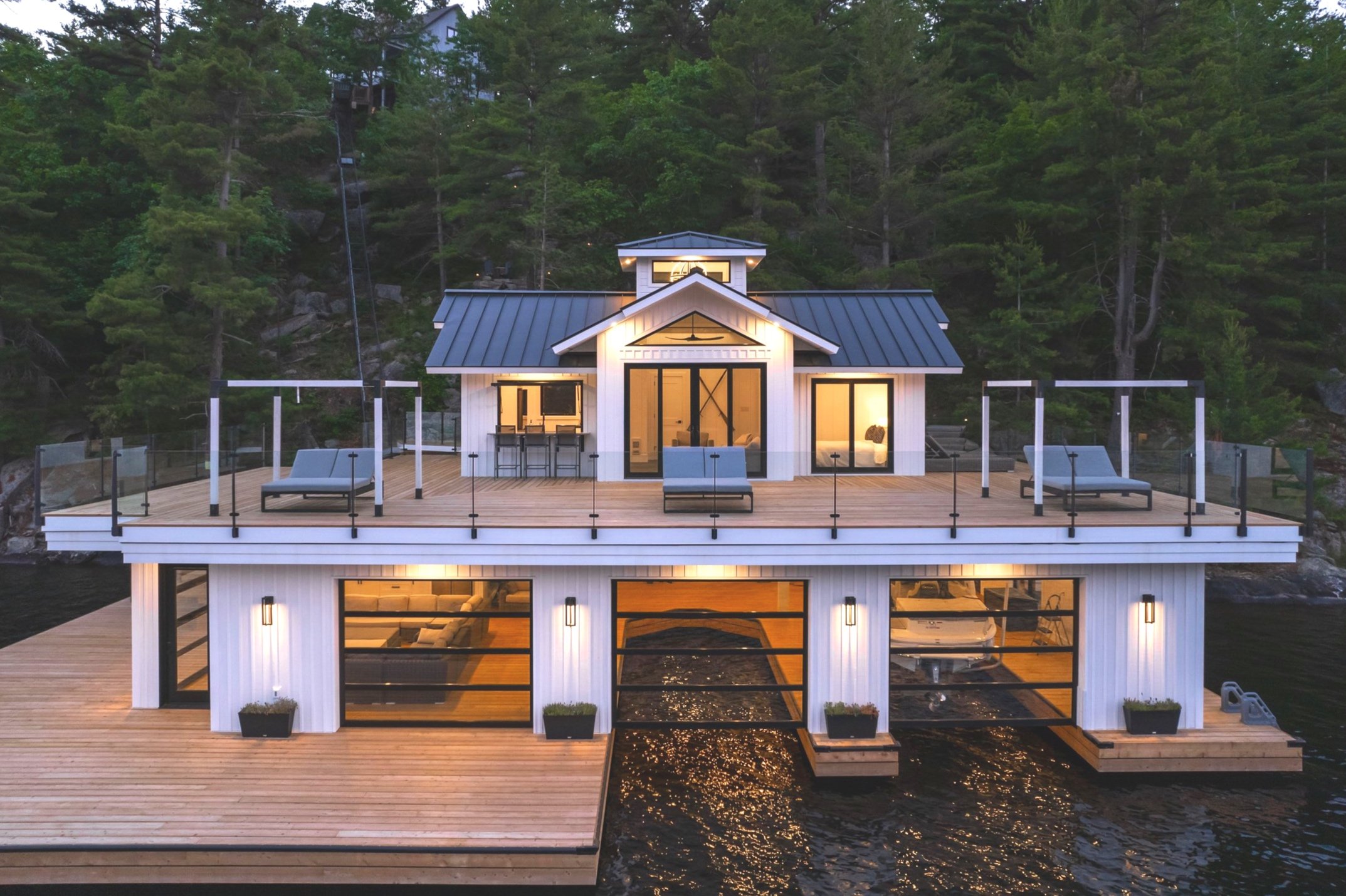 A modern two-story house on a waterfront with large glass windows, a spacious wooden deck, and surrounding lush green trees.