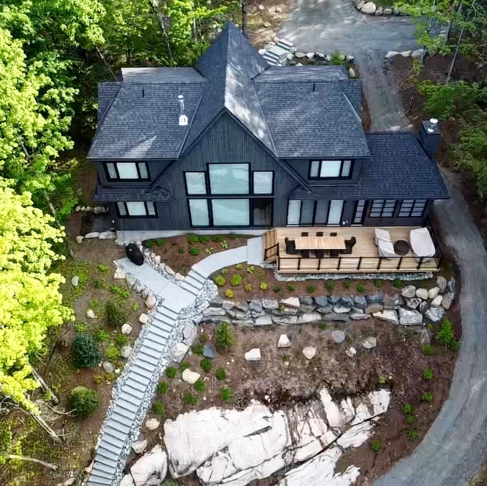 Aerial view of a modern black house with large windows, a balcony with outdoor furniture, surrounded by lush green trees and a curved driveway.