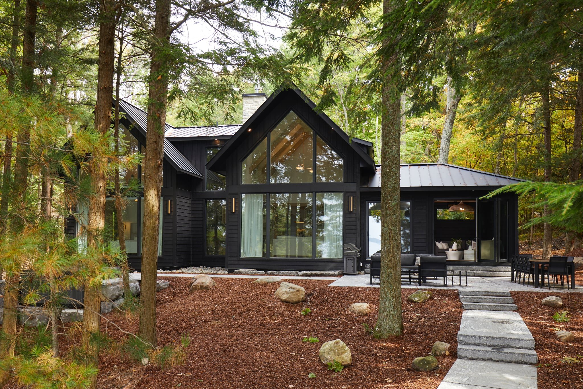 Black modern house with large glass front, surrounded by trees in a forest setting, with stone steps leading to a patio area.