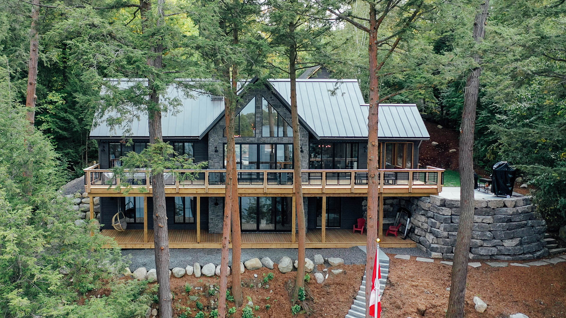 A modern, multi-story house with large glass windows and a wooden deck, situated among tall trees in a forested area.