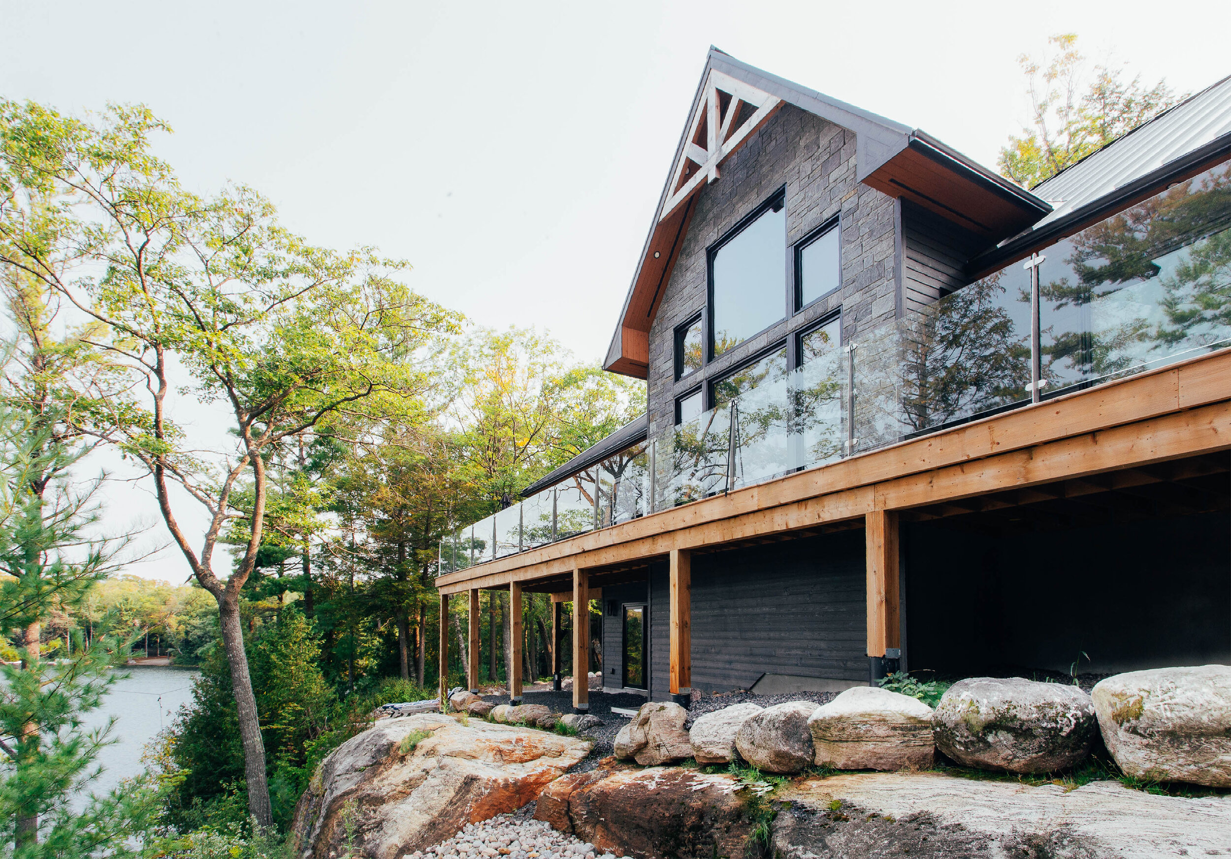 Modern house with large glass windows and wooden deck, built on a rocky hillside near trees and water
