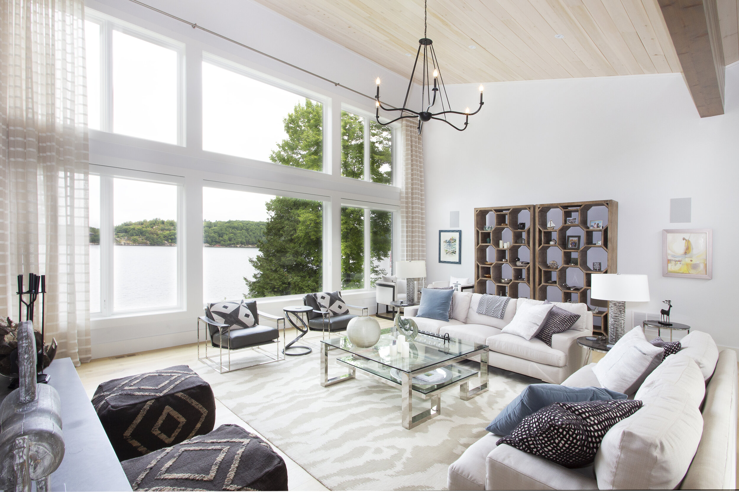 Bright living room with large windows overlooking a lake, featuring white sofas, black and patterned accent chairs, a glass coffee table, and wooden shelving, with a high ceiling and chandelier.