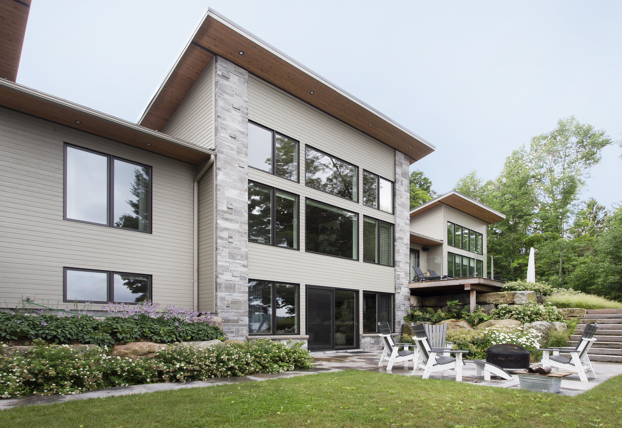 Modern multi-story house with large windows, stone and siding exterior, surrounded by greenery and a landscaped yard with seating area, lawn, bushes, and trees.