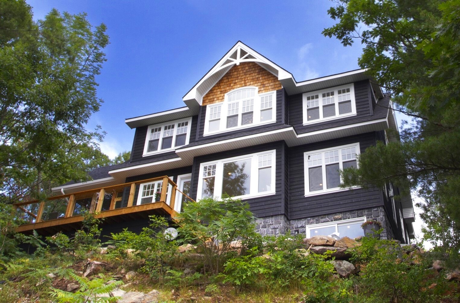 A large, multi-story house with black siding, white trim, and a stone foundation, surrounded by greenery and trees.