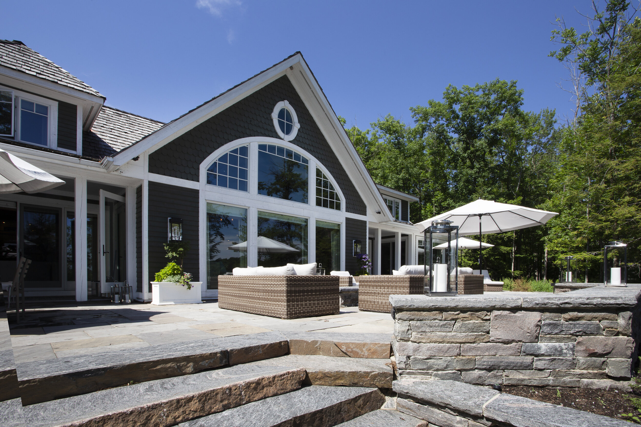 Backyard patio of a house with wicker furniture, umbrellas, and lanterns, surrounded by trees.