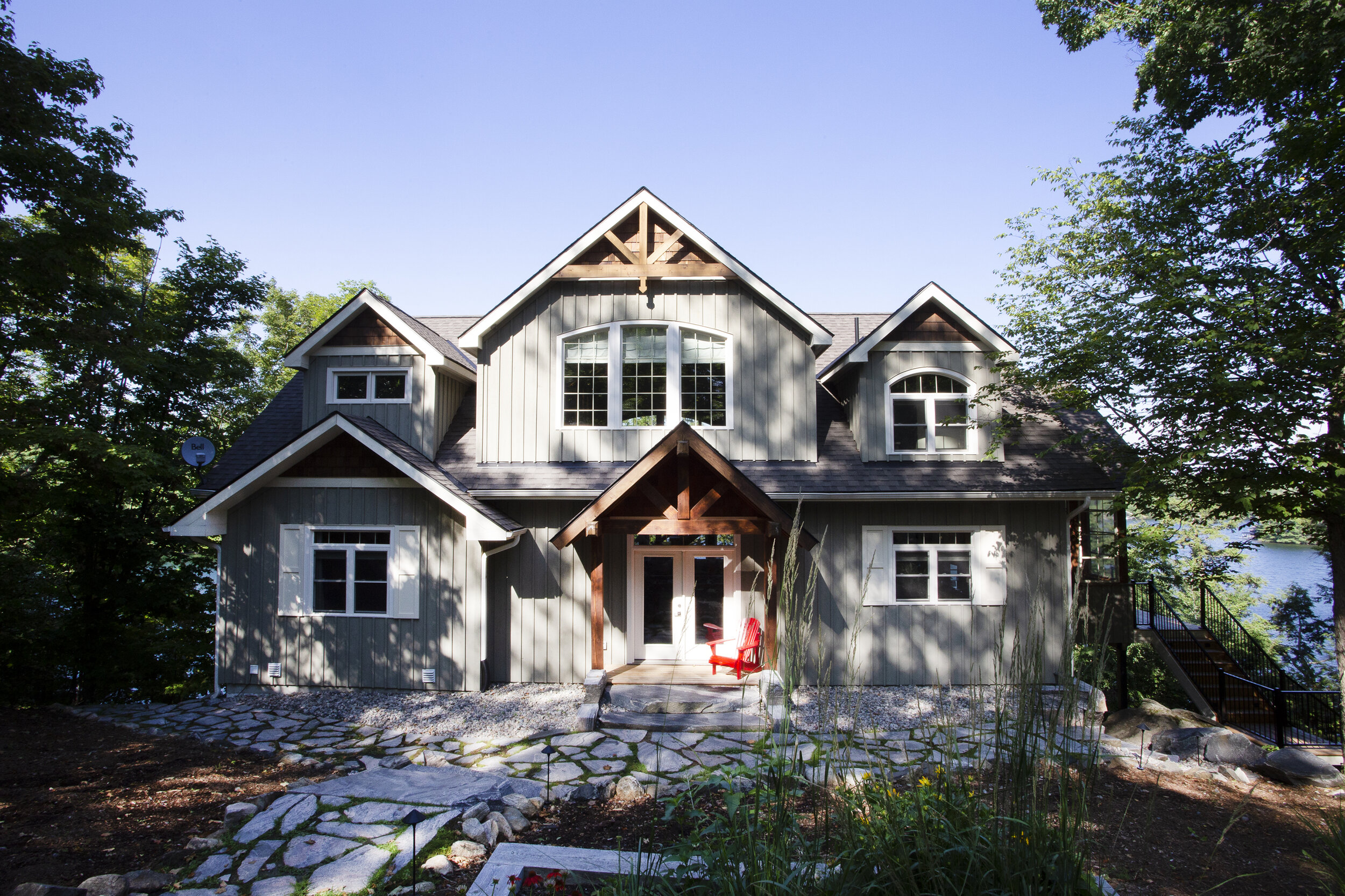 A modern, gray multi-story house with a front porch, gabled roof, and several arched and rectangular windows, surrounded by trees in a natural setting.