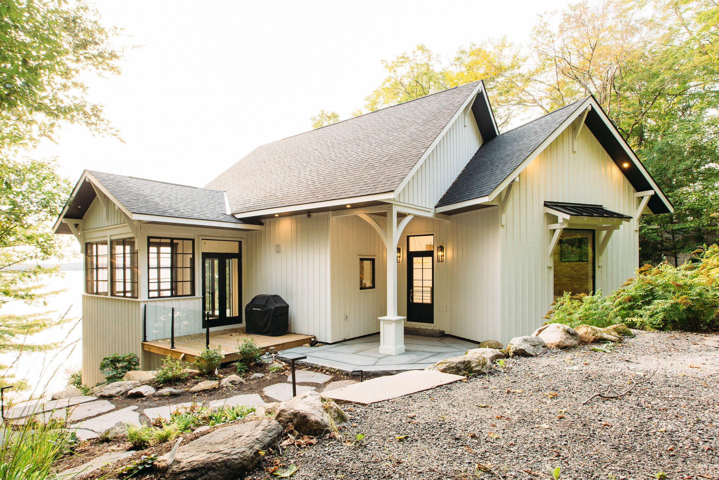 A modern house with white vertical siding, black-framed windows and doors, a small deck with a charcoal grill, amid trees with autumn leaves.