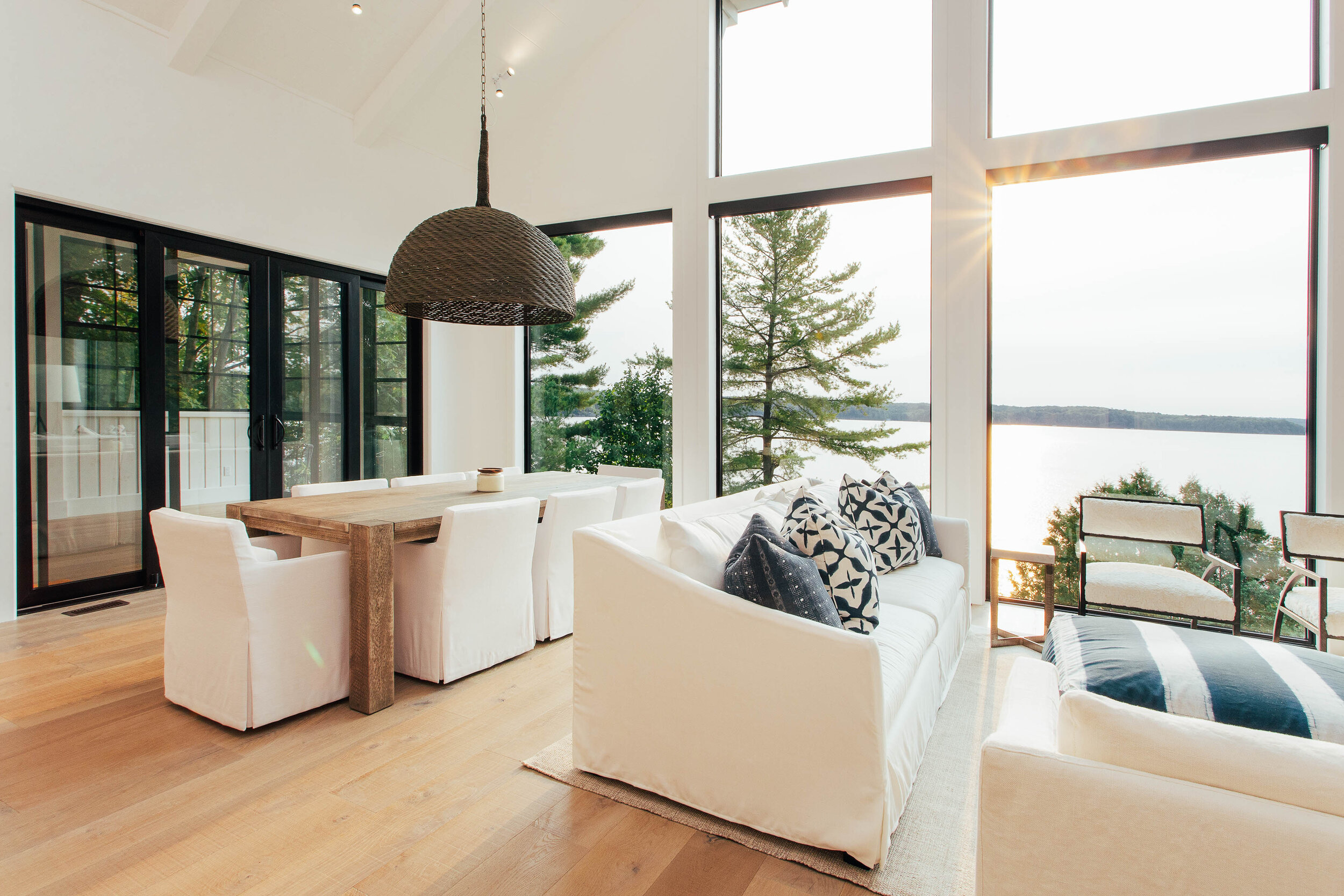 Living room with large windows overlooking a lake, featuring a white sofa with black and white patterned pillows, a wooden dining table with white chairs, and two armchairs with a bench outside on a balcony.