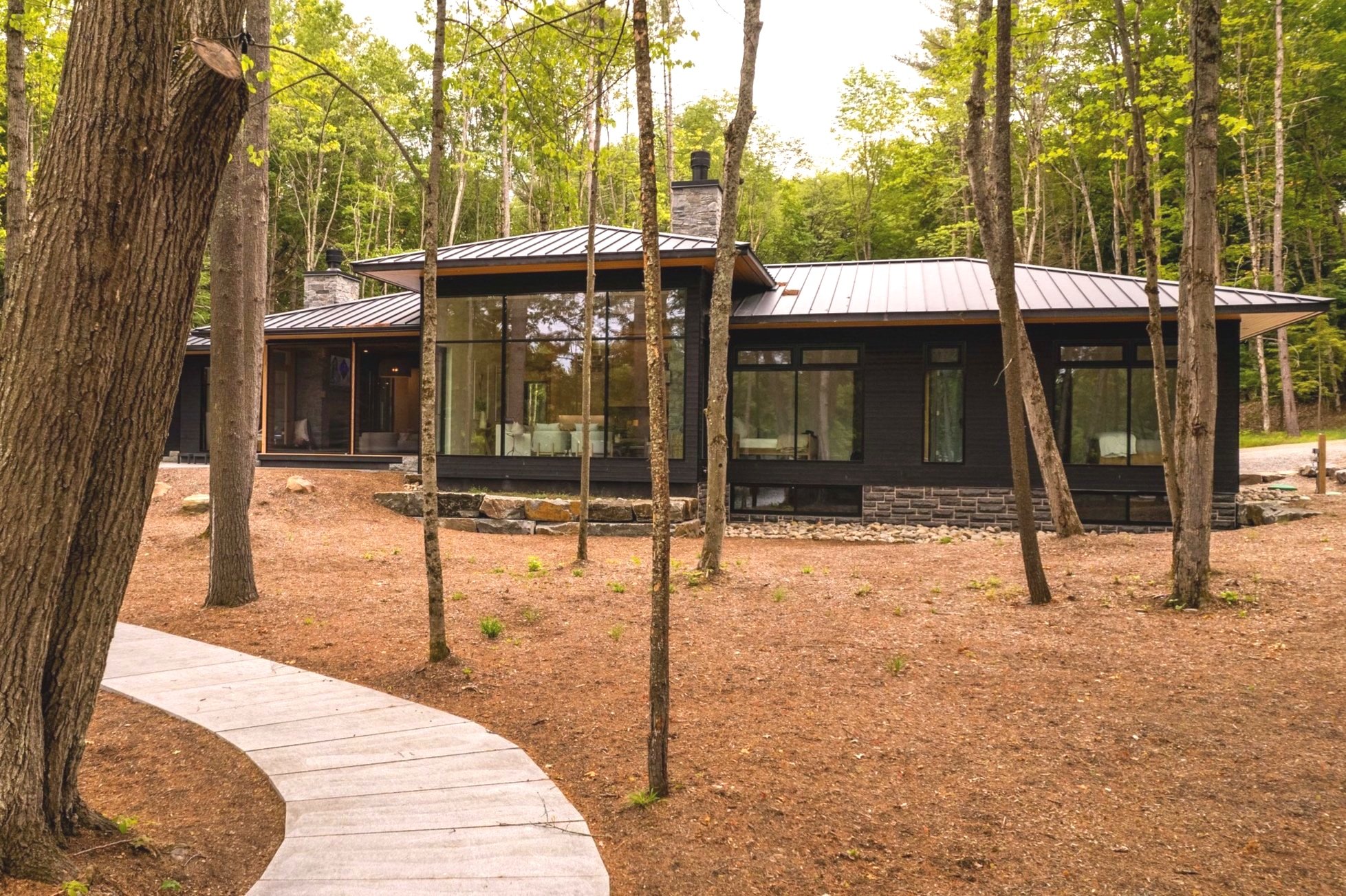 Modern house with large glass windows surrounded by trees, a curved walkway in the front, and a wooded area in the background.