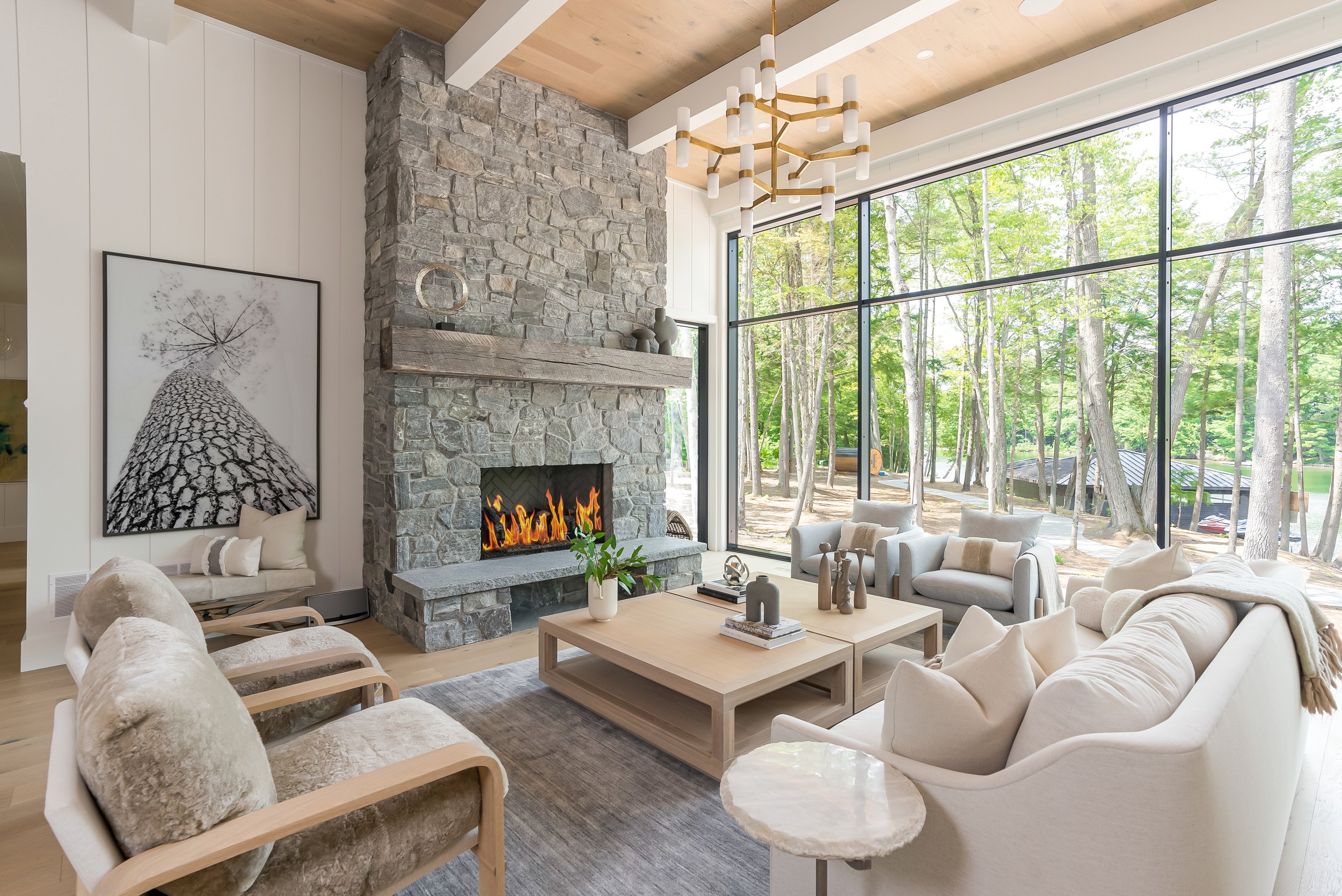 Living room with stone fireplace, large windows overlooking trees, white and beige seating, wooden coffee table, modern chandelier, and artwork on the wall.