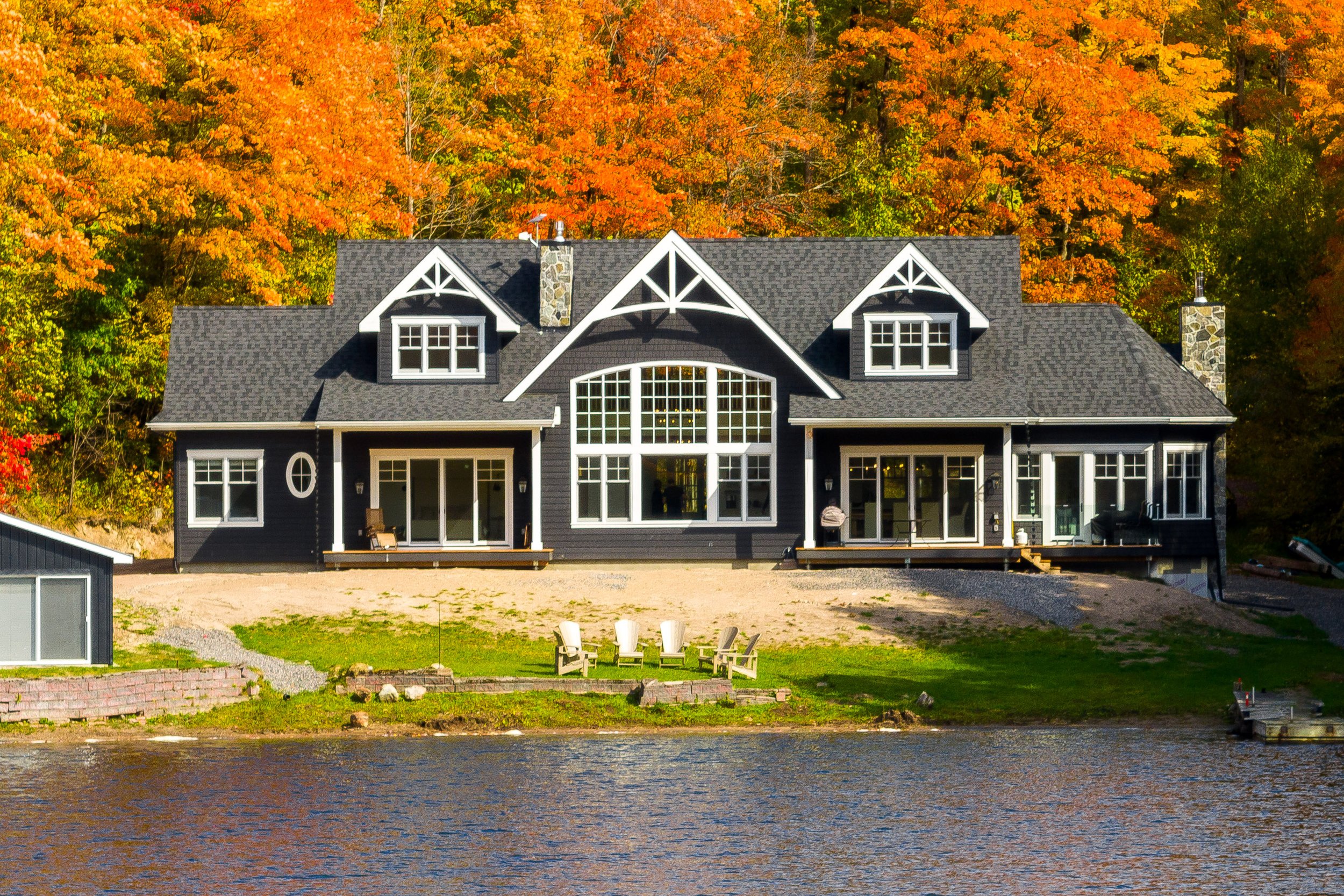 Large black house with multiple windows and a steep roof, situated near a body of water with a lawn and chairs in the foreground, surrounded by colorful autumn trees.