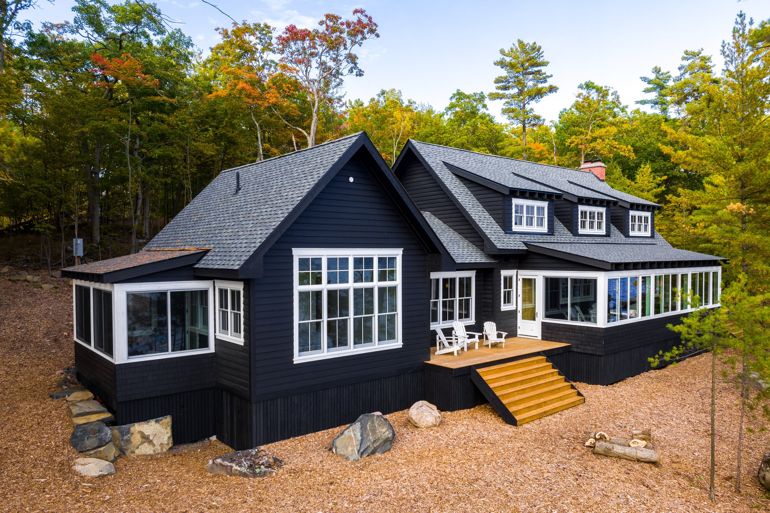 A black house with large windows, a wooden front porch, and surrounding trees with autumn foliage.