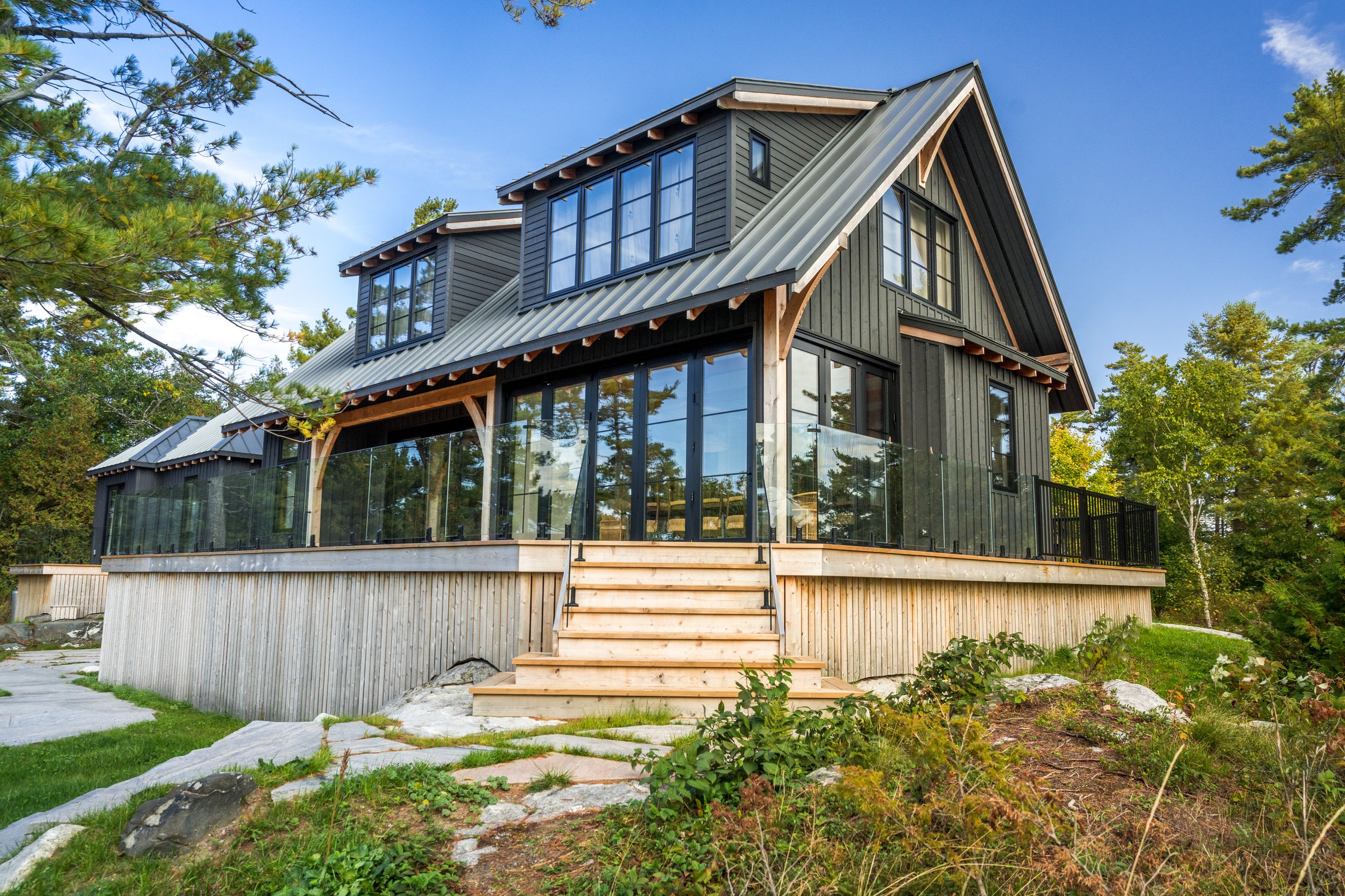 A modern, multi-story house with black exterior siding and large windows, surrounded by trees and greenery, with a wooden staircase leading to a glass-enclosed porch.