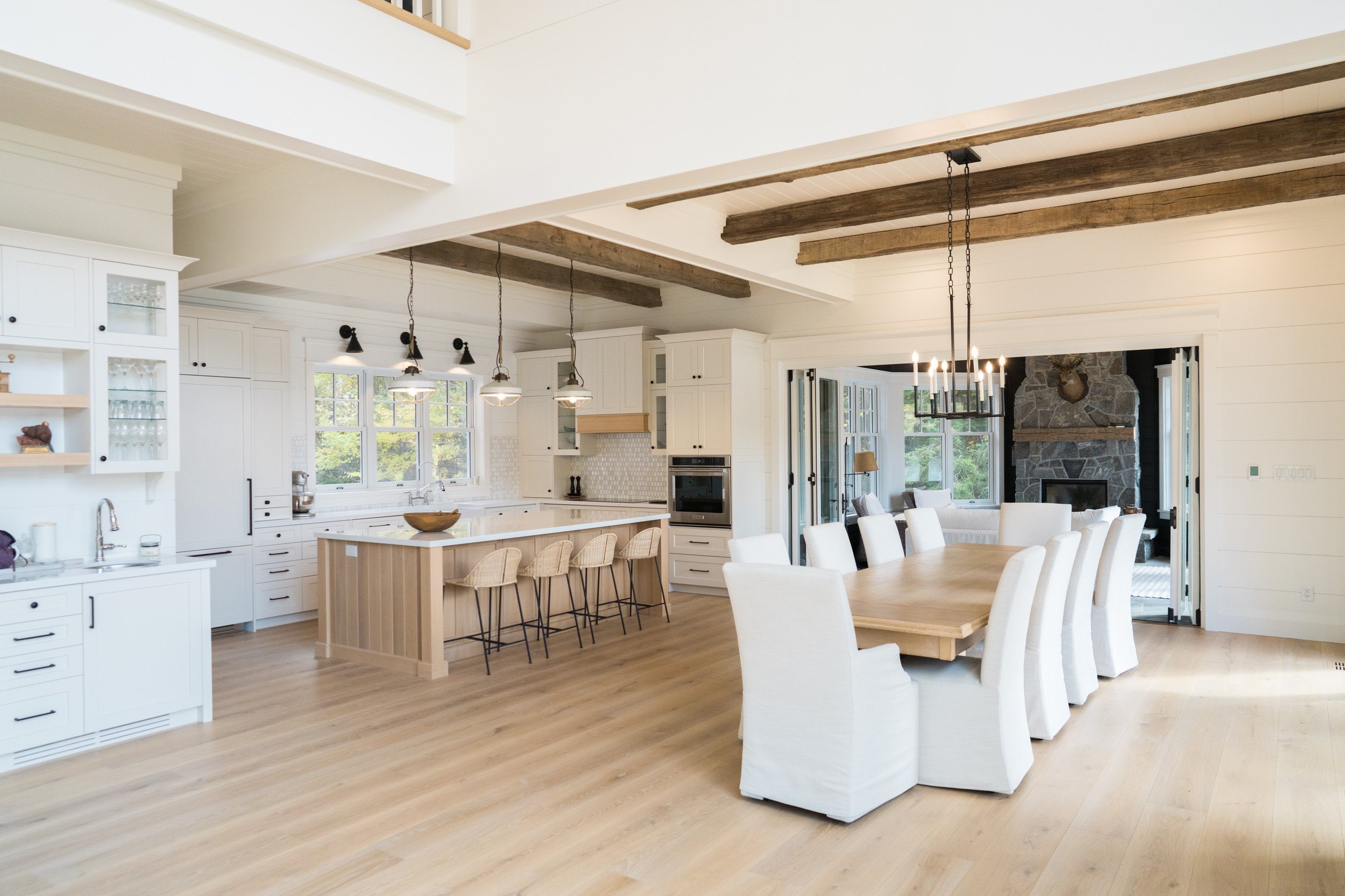 Bright open-concept kitchen and dining area with white cabinetry, wooden beams, and a stone fireplace in the background.