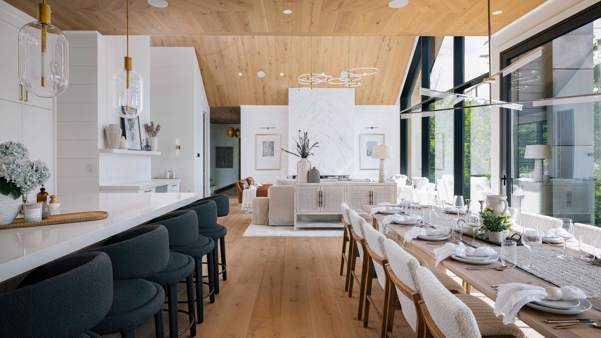 Bright open dining and living space with large windows, wooden ceiling, and modern decor. Dining table set with white plates, glasses, and napkins; living area has a beige sofa, coffee table, and abstract artwork.