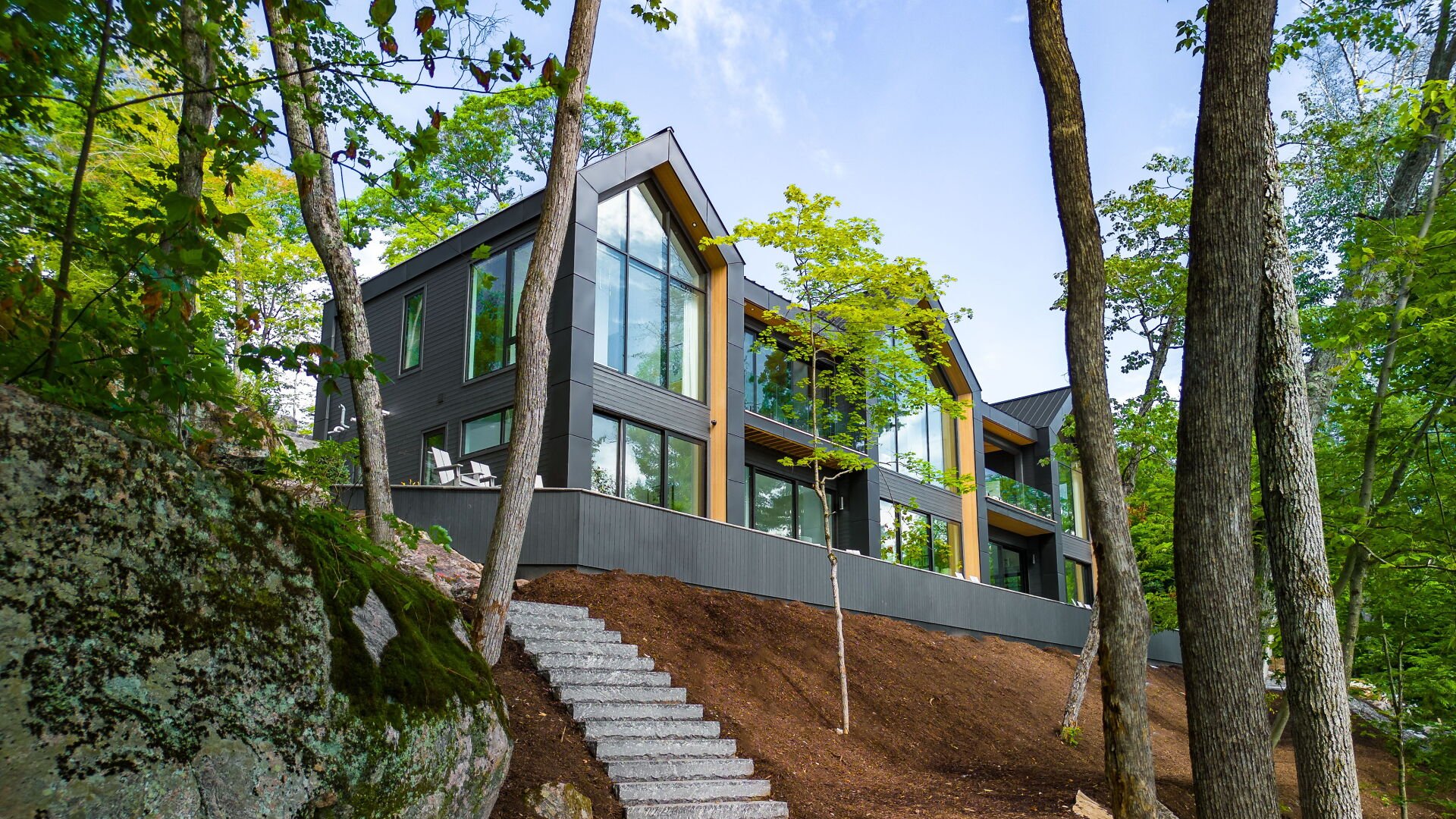 Modern multi-story house with large glass windows on a hillside surrounded by trees and rocky terrain.