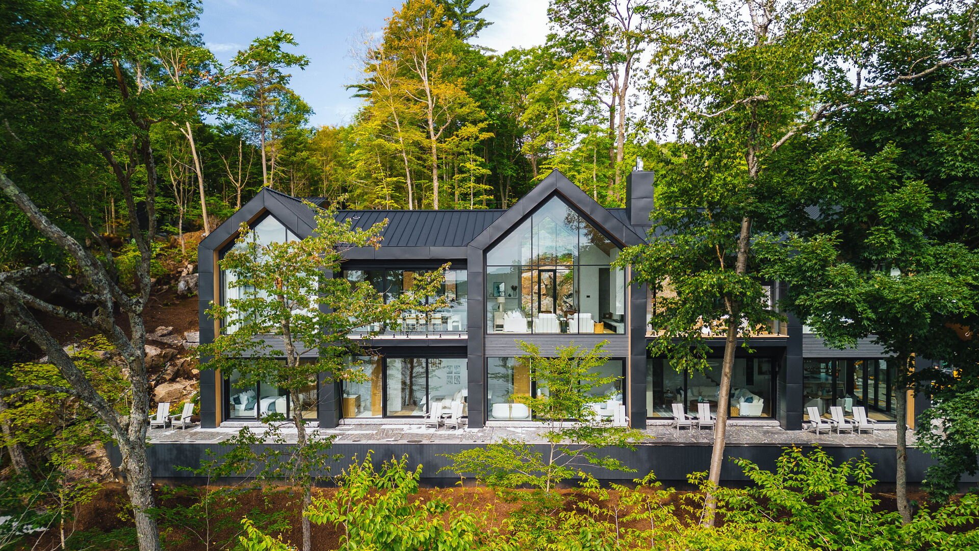 Modern two-story house with large glass windows surrounded by trees, with a balcony and outdoor seating area.